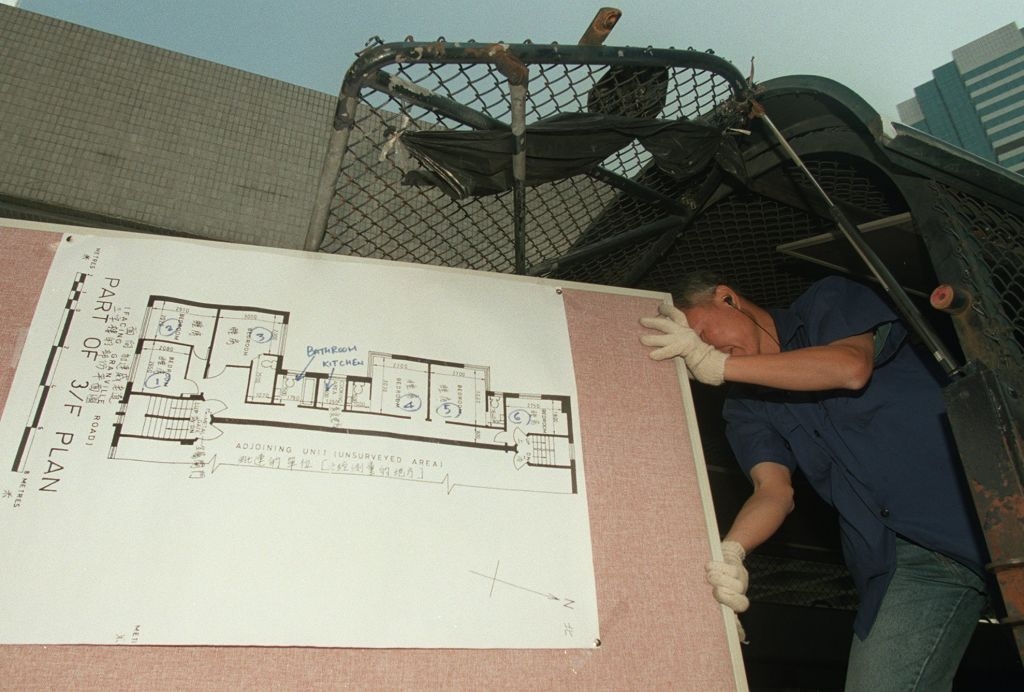 Person i blå skjorte og hansker flytter en stor arkitektonisk planplate utenfor en bygning. Tavlen viser en detaljert planløsning