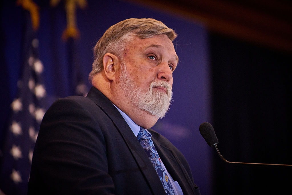 En person med skjegg snakker ved et podium med flagg i bakgrunnen, iført dress og mønstret slips