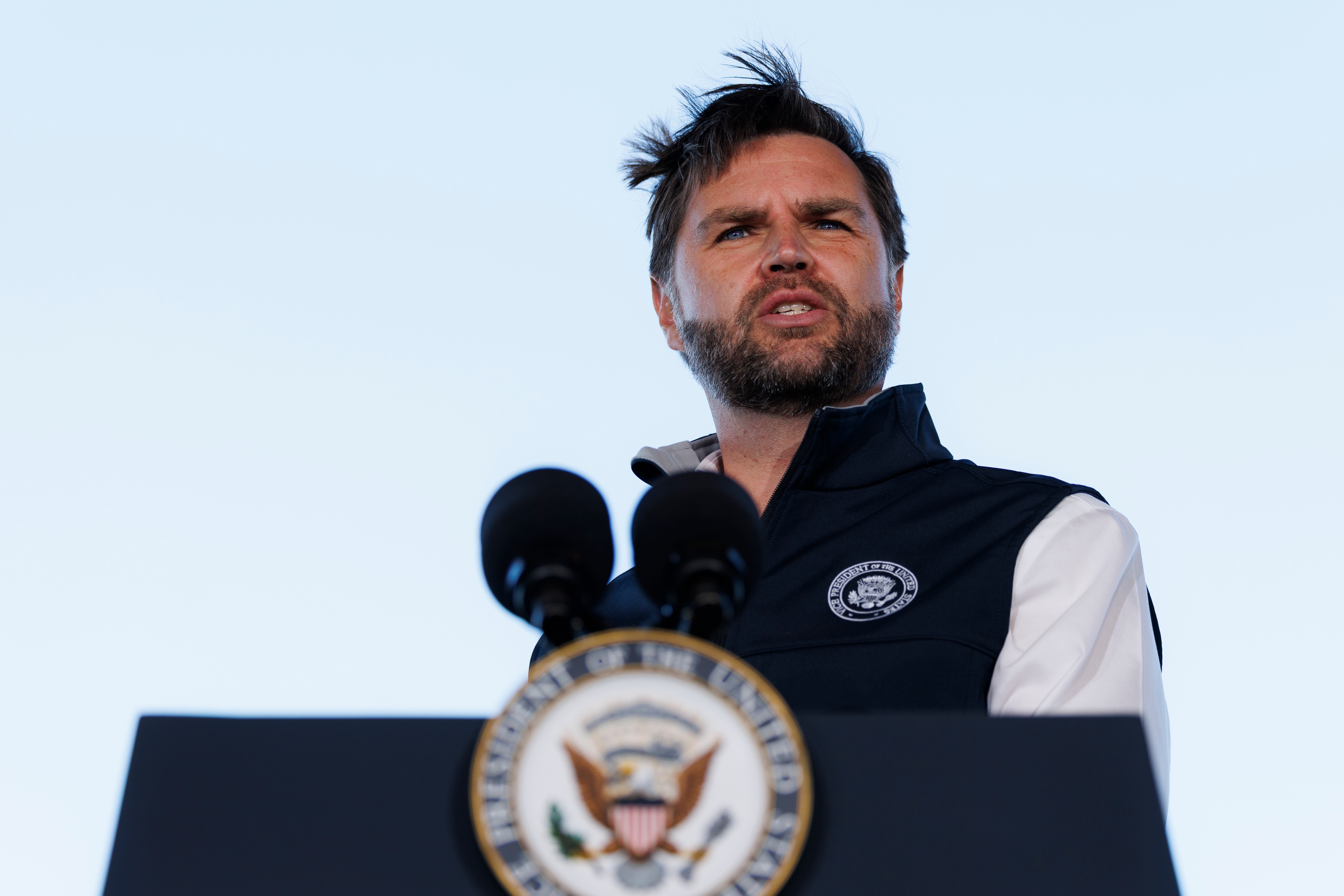 En mann står på et podium med en regjeringssegl, snakker inn i flere mikrofoner, iført en ermeløs jakke over en hvit skjorte