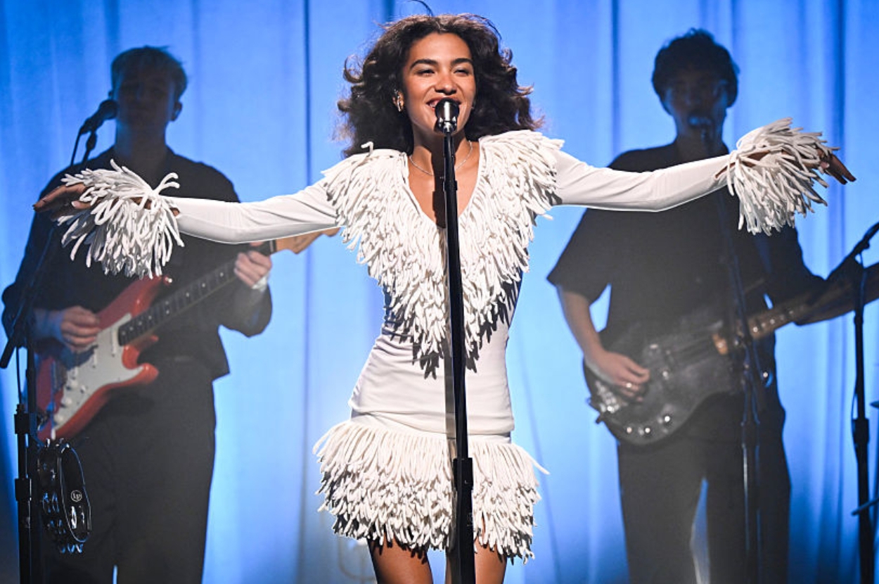 Singer performs on stage in a fringed dress, arms outstretched, accompanied by a band in the background