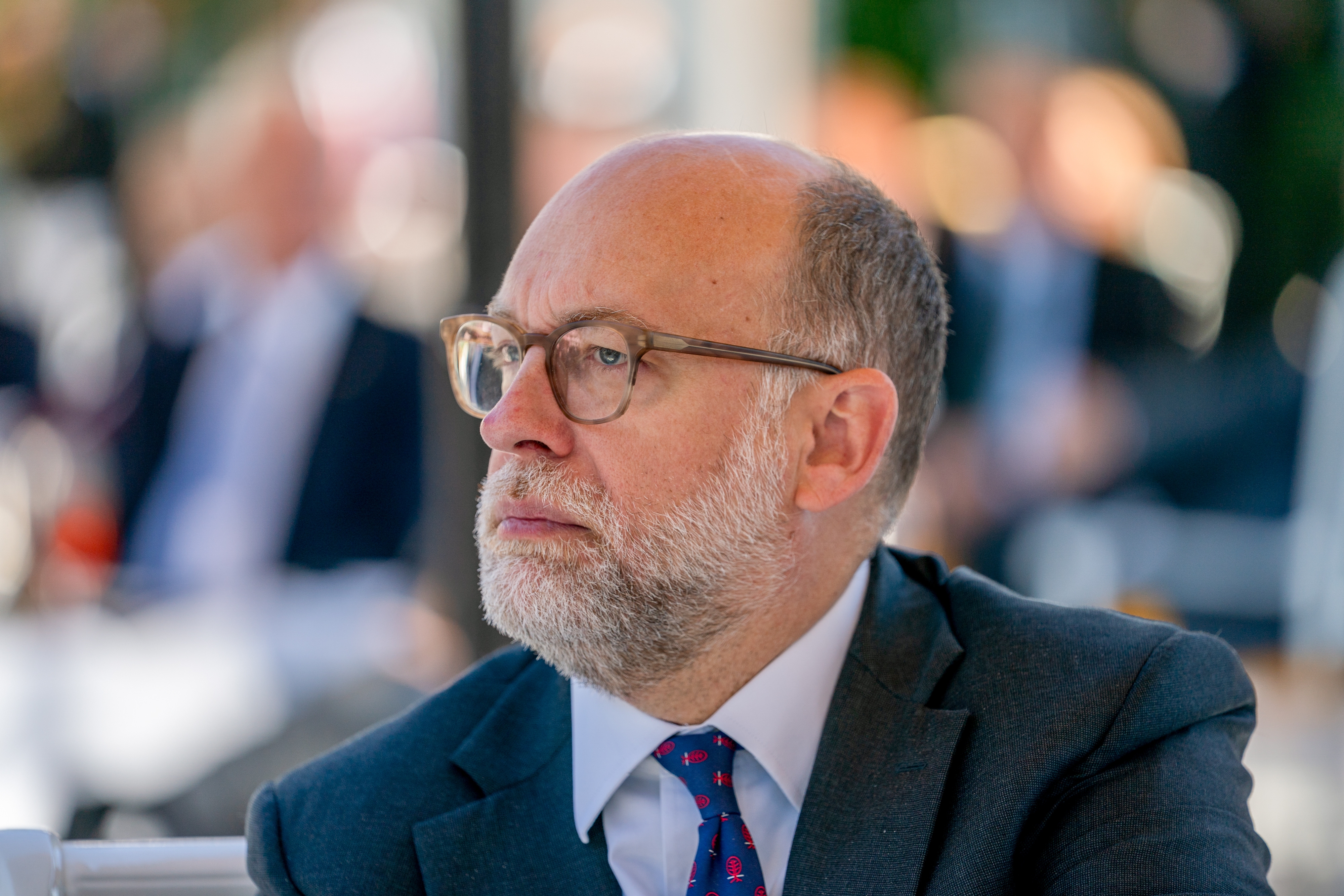 En mann med briller, dress og slips med et gjennomtenkt uttrykk på et utendørsarrangement