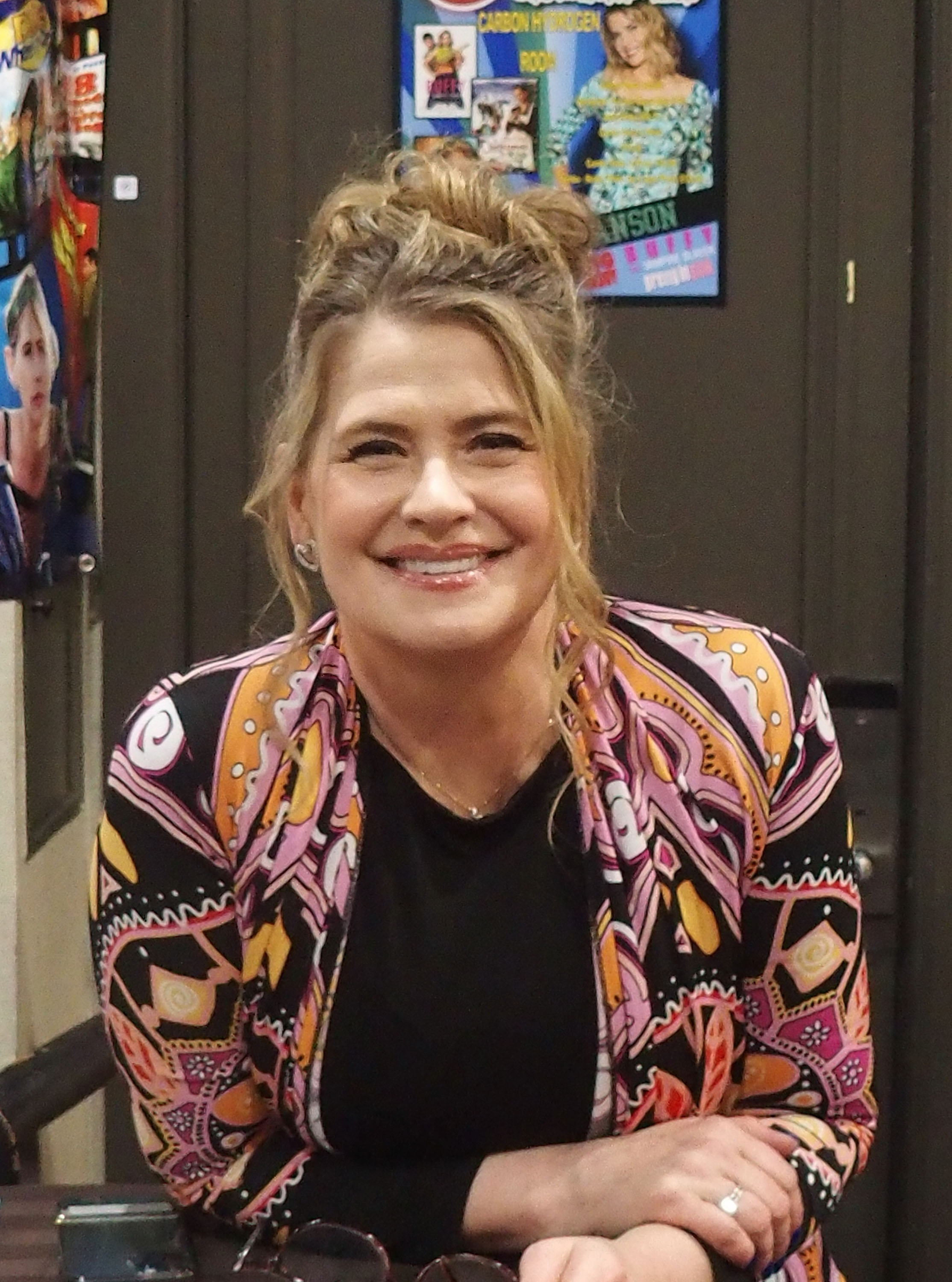 Person som smiler på et arrangement, iført en mønstret jakke med geometriske design. Plakater er synlige i bakgrunnen