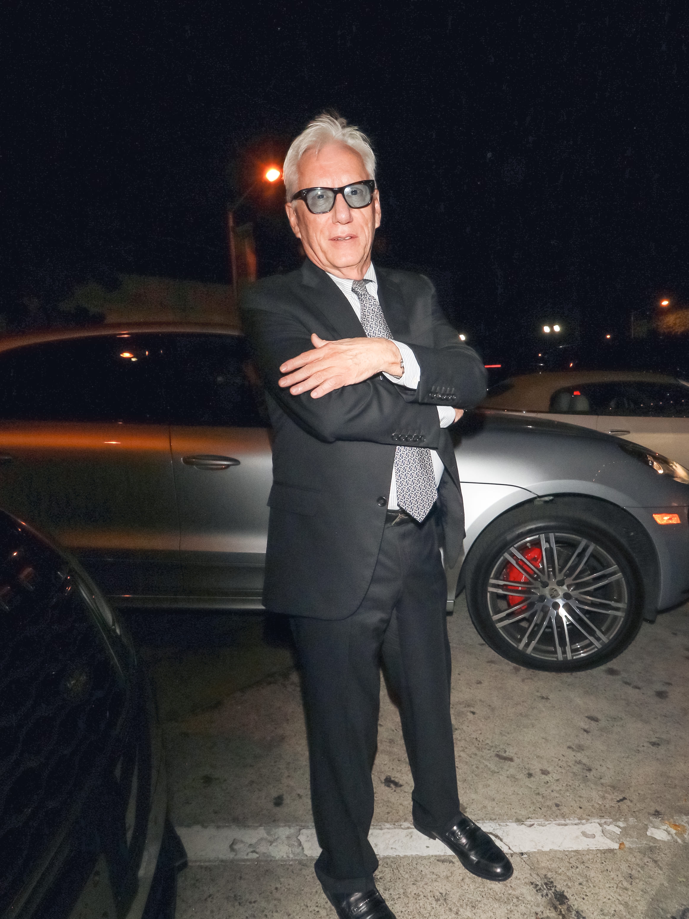 Mann i dress og slips med solbriller poserer selvsikkert på en parkeringsplass om natten