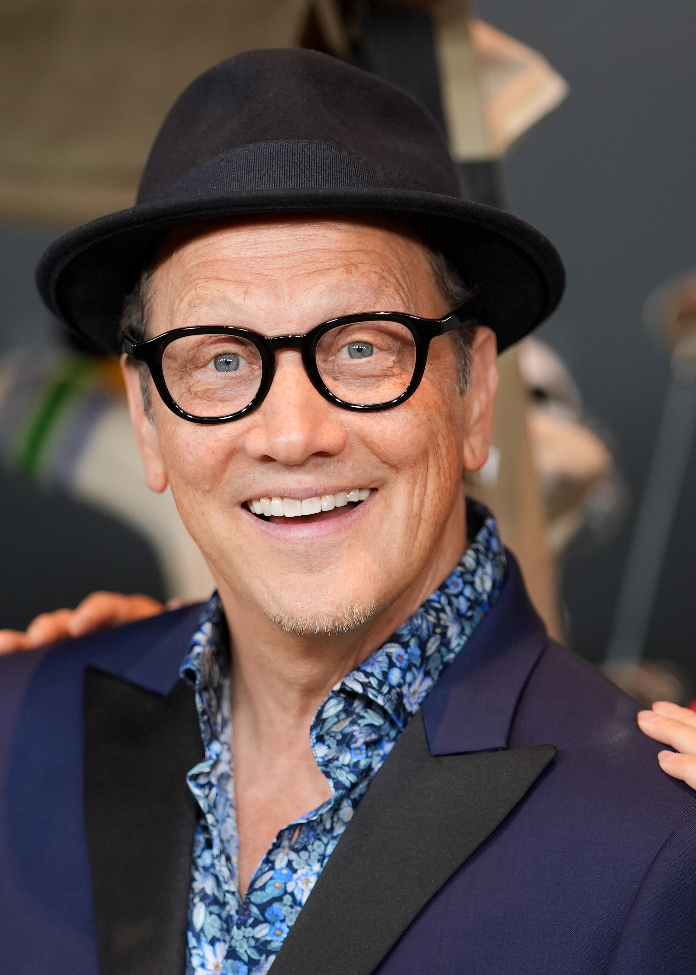 En person smiler til kameraet, iført en hatt, briller og en blomsterskjorte under en dressjakke, på et kjendisarrangement