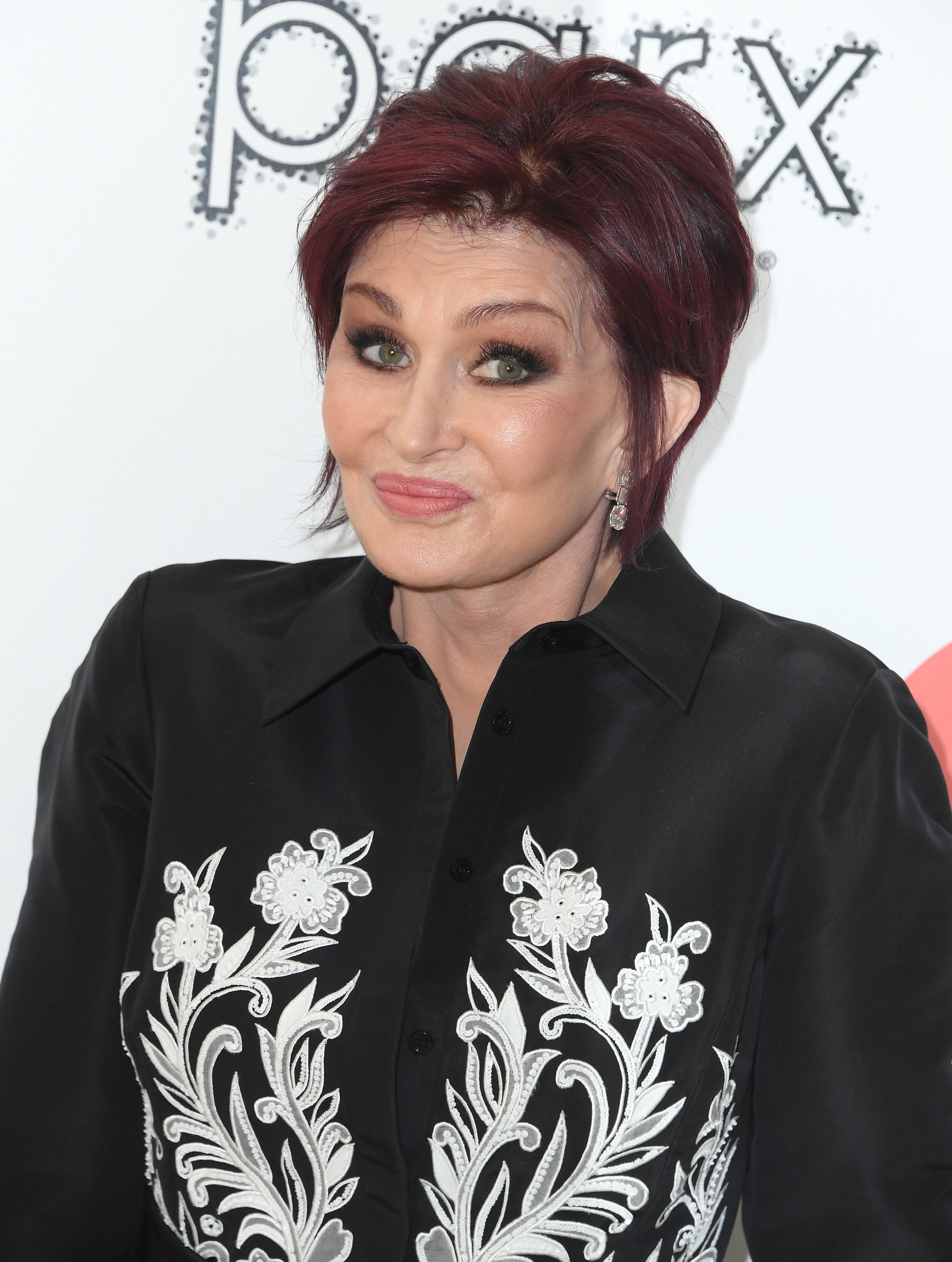 Sharon Osbourne posing, wearing a achromatic  blouse with intricate achromatic  floral embroidery