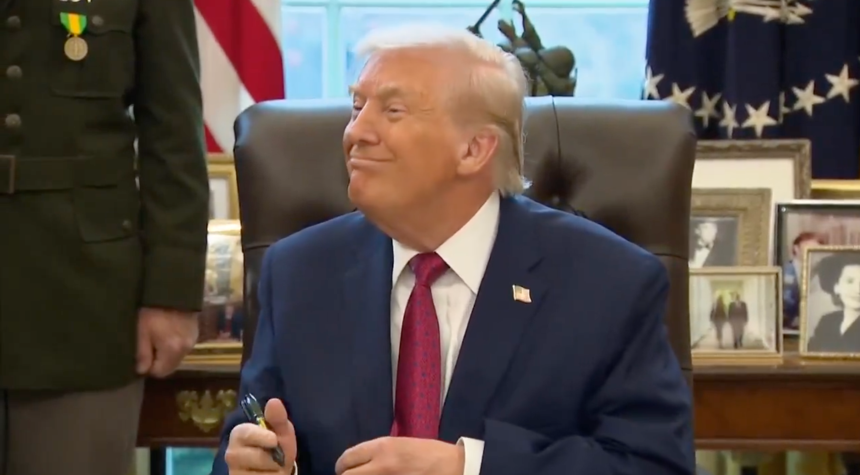 A idiosyncratic successful a suit sits astatine a table with a pen, smiling, surrounded by photos and awards successful an bureau setting