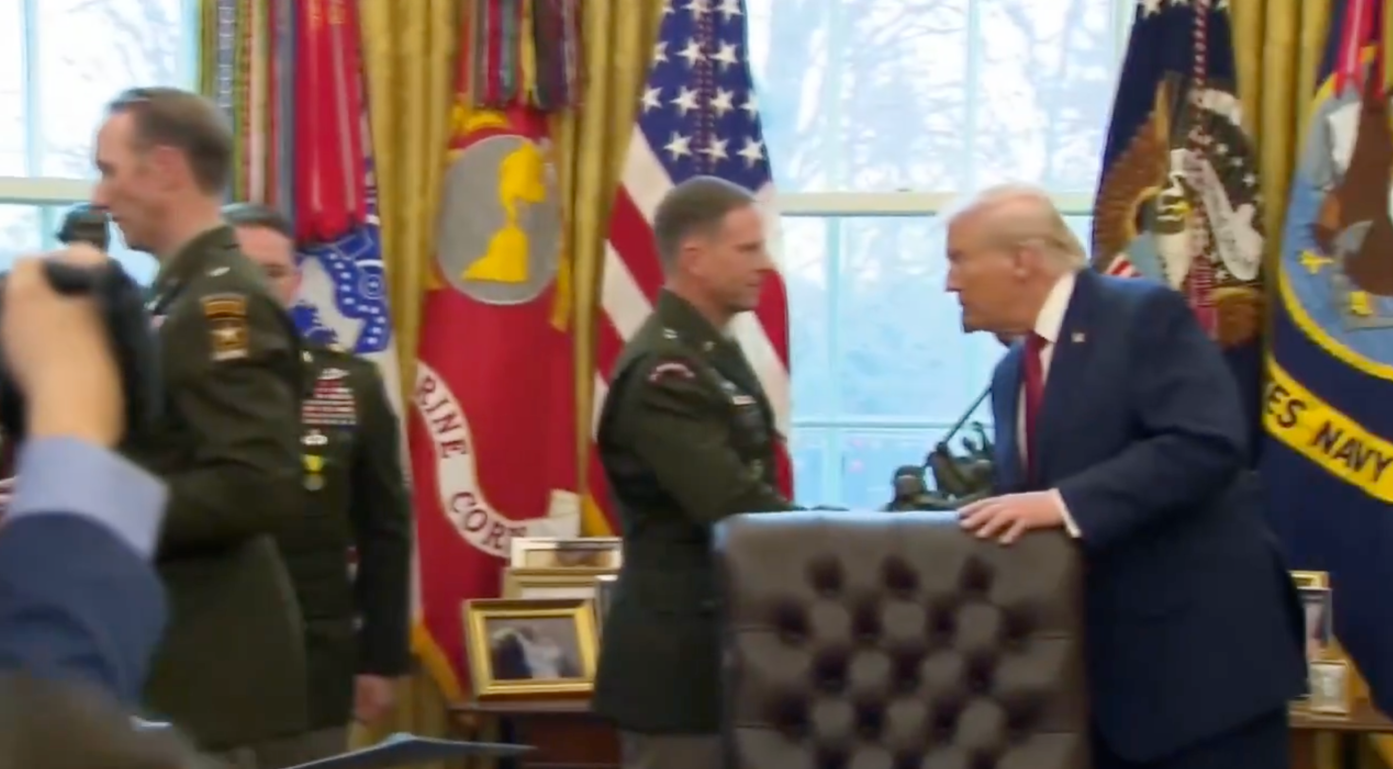 A antheral successful a suit shakes hands with a uniformed subject serviceman successful an authoritative country with flags successful the background