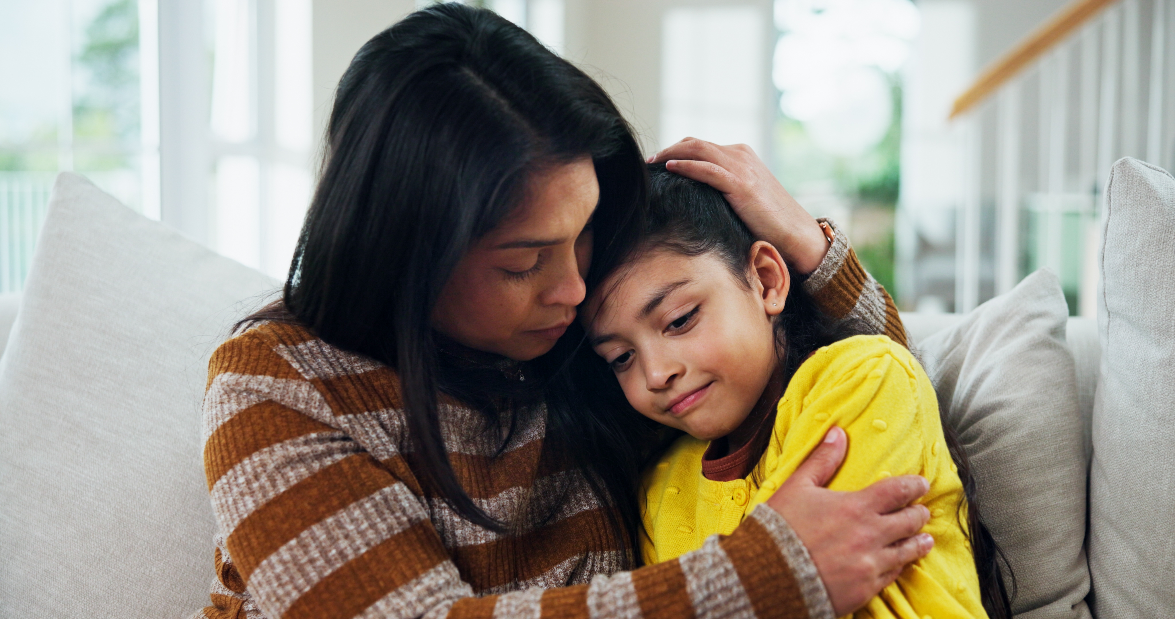 A pistillate mildly hugs a kid connected a couch, offering comfortableness and support. The kid rests their caput connected the woman's shoulder, looking content