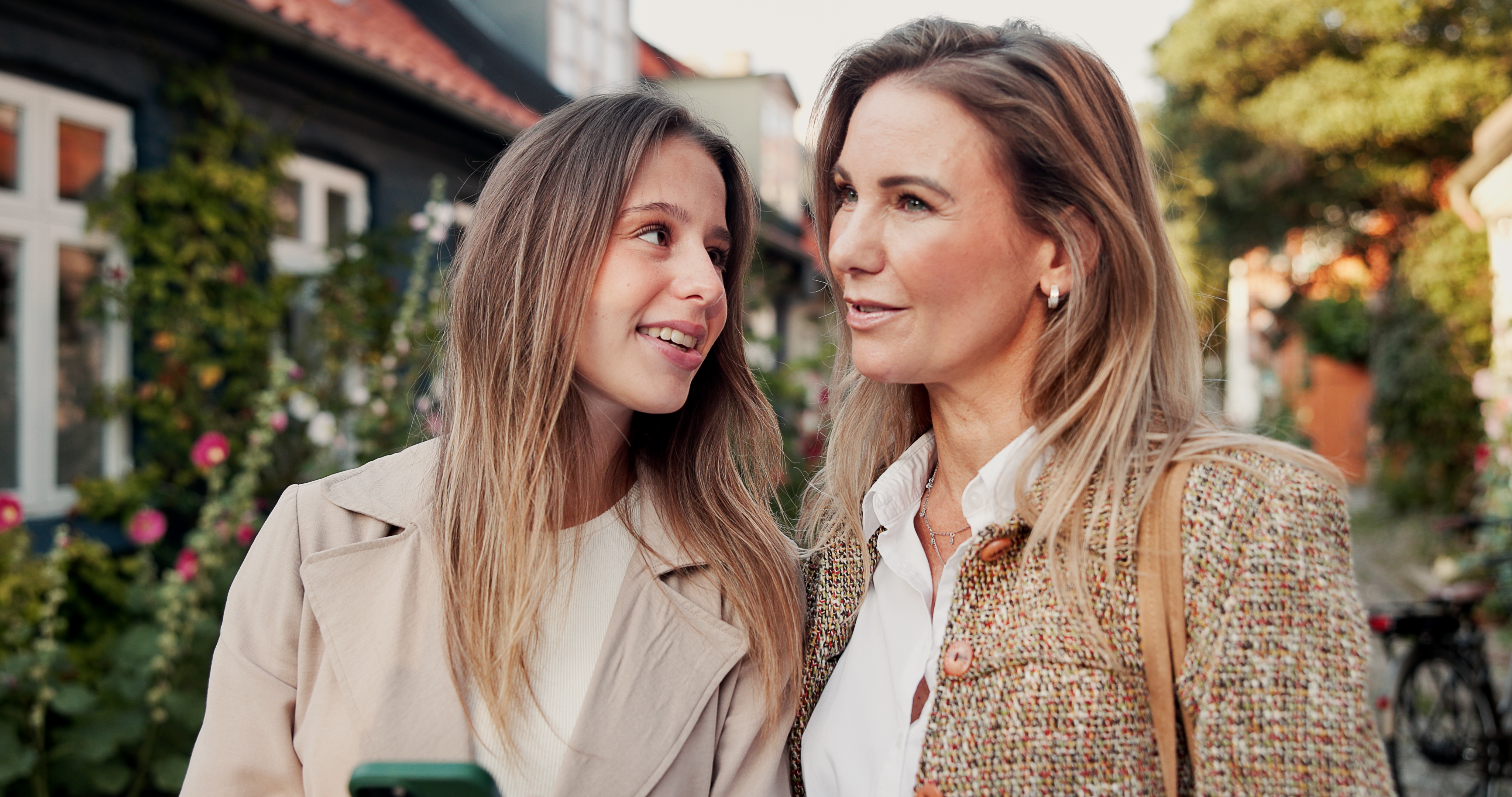 Two people, a younger and an older person, smiling astatine each different portion walking connected a charming thoroughfare lined with flowers and houses