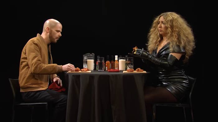 Two people at a table with hot sauces and milk, one in a brown jacket and the other in a black leather dress, appear to be on a talk show