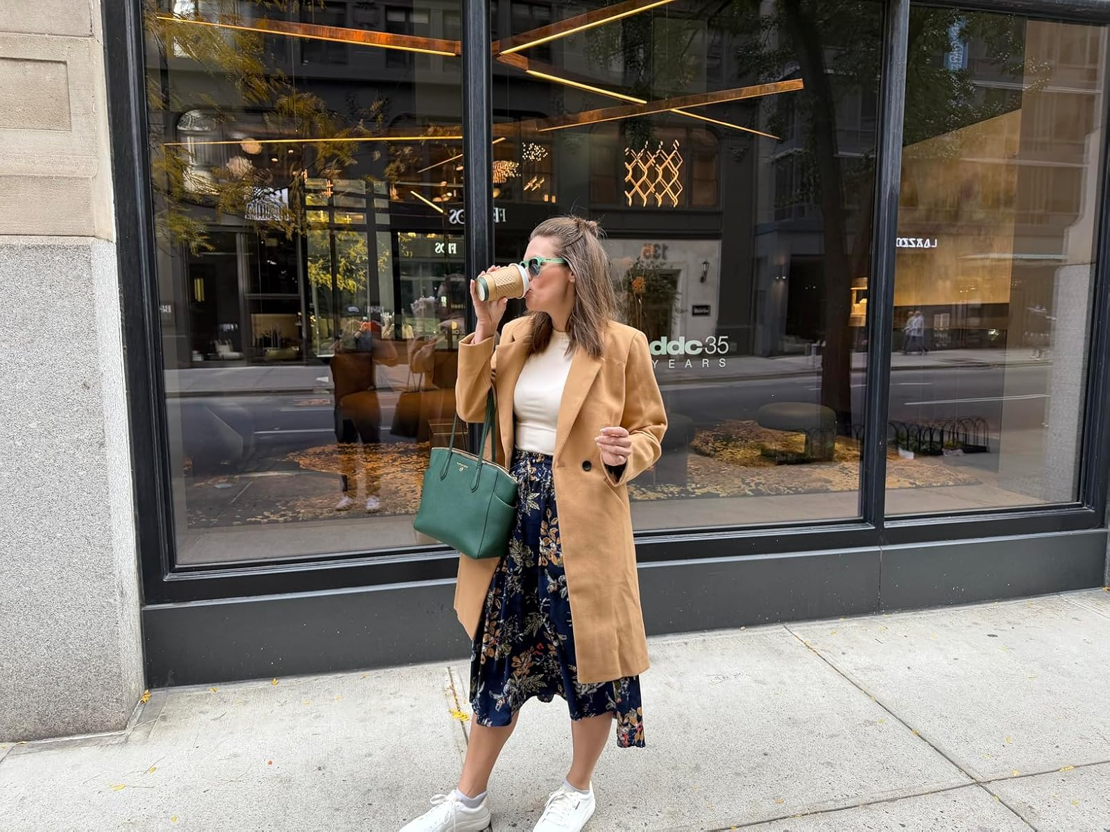 Person successful  a agelong  camel coat, floral skirt, and sneakers sips coffee, holding a greenish  bag, extracurricular  a stylish storefront