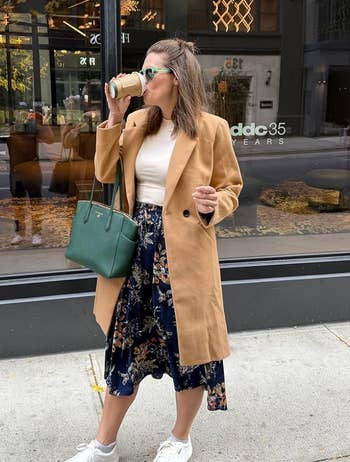 Person successful a agelong camel coat, floral skirt, and sneakers sips coffee, holding a greenish bag, extracurricular a stylish storefront