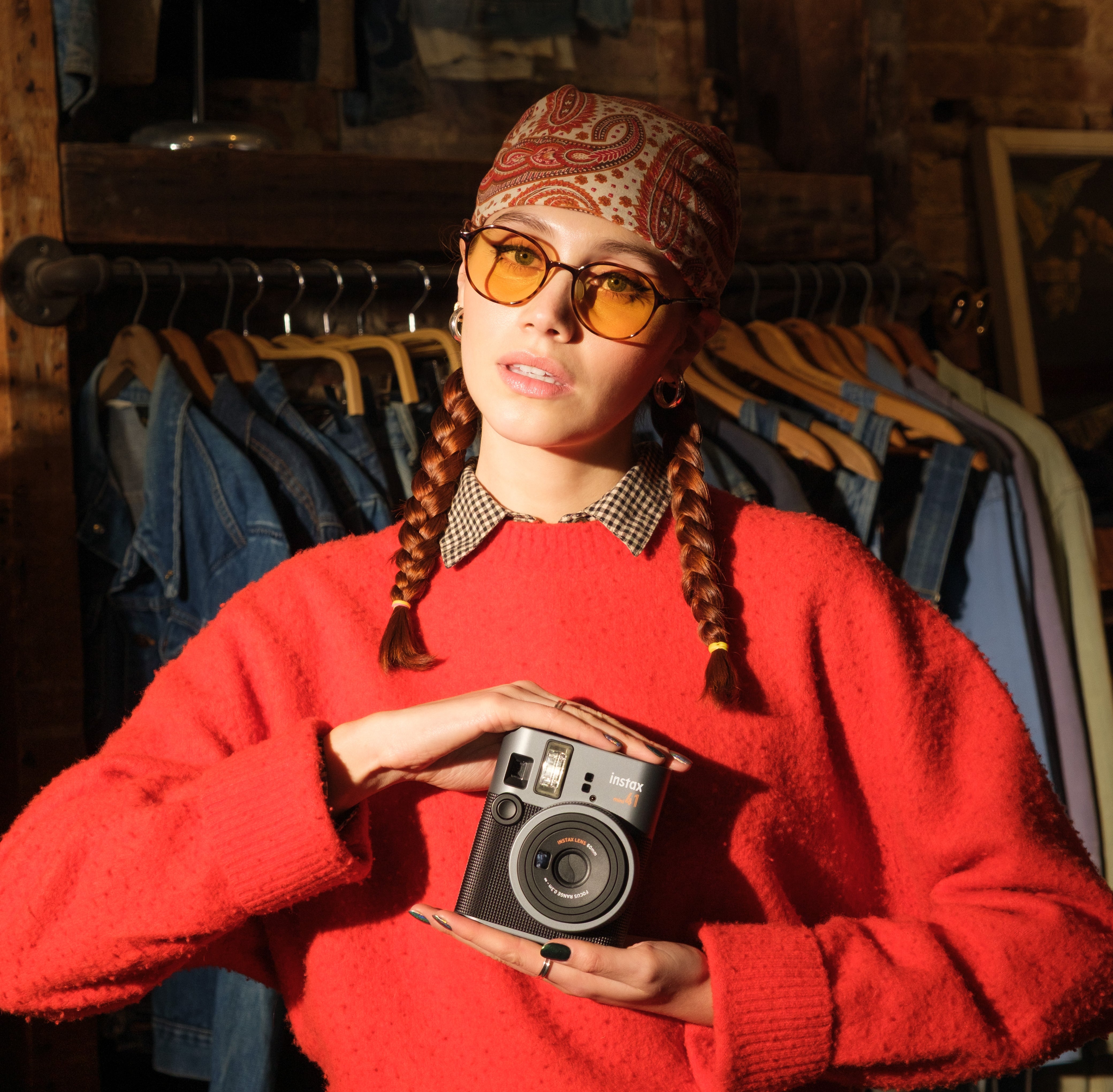 Person in a cozy sweater and patterned headscarf holds a camera, standing in front of vintage clothing racks