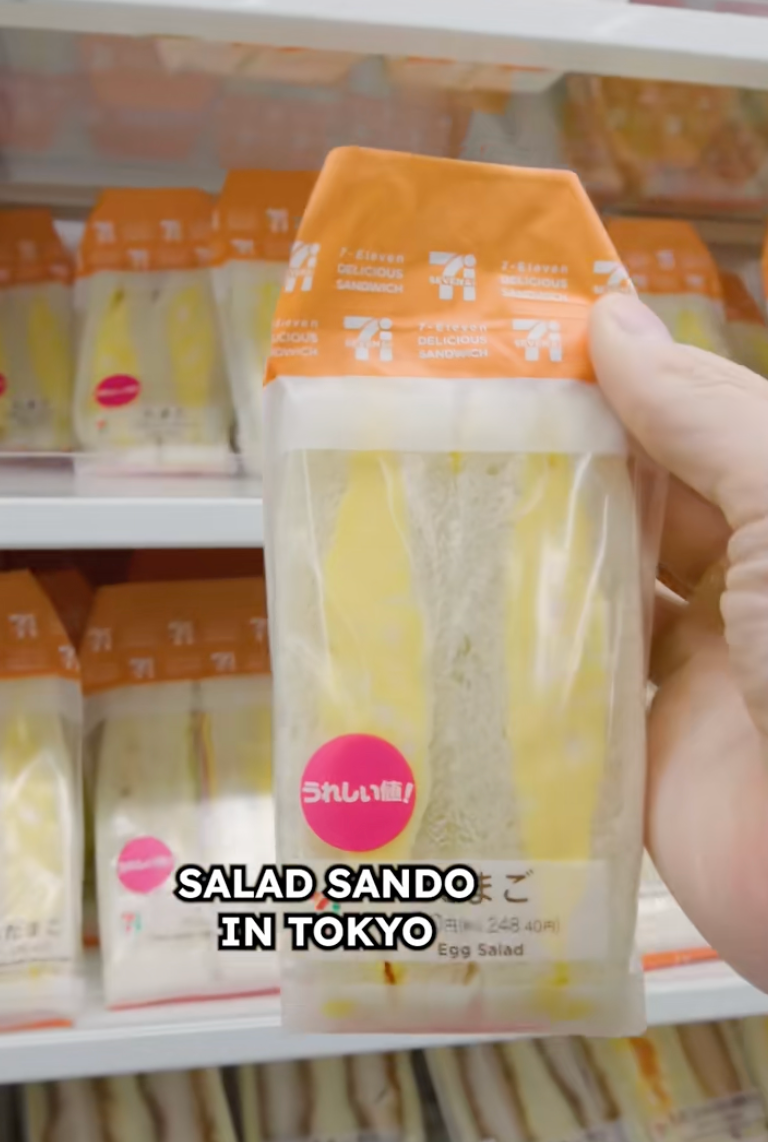 Hand holding a packaged egg salad sandwich in a Tokyo convenience store. Nearby shelves display similar sandwiches. Text reads "Salad Sando in Tokyo."