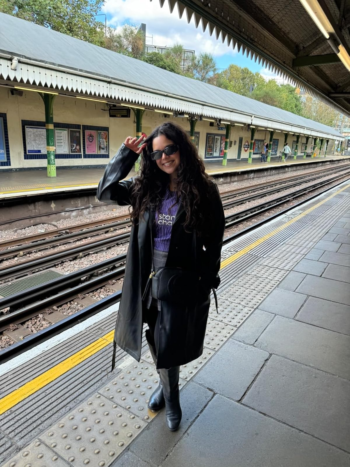 Person smiling astatine  a bid     presumption    wearing a agelong  coat, sunglasses, and boots, with a manus  raised somewhat  holding onto eyewear