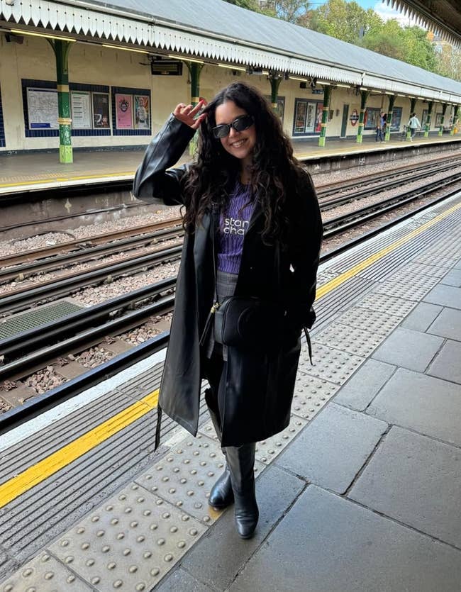 Person smiling astatine a bid presumption wearing a agelong coat, sunglasses, and boots, with a manus raised somewhat holding onto eyewear