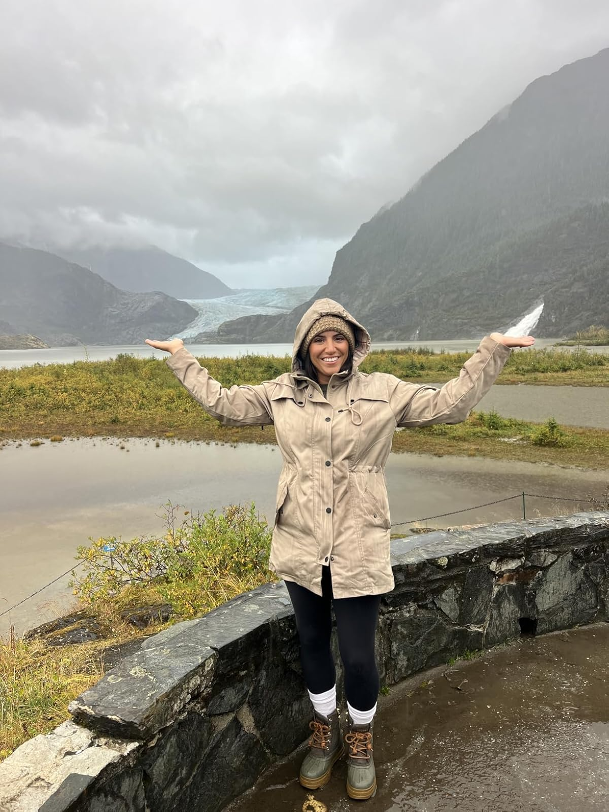 A idiosyncratic   successful  a hooded overgarment   smiles by a chromatic  railing, with mountains and a distant   glacier successful  the background, against a cloudy sky