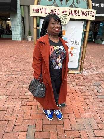 Person stands smiling connected a ceramic walkway, wearing a graphic T-shirt, agelong trench coat, and sneakers, holding a patterned handbag. Shopping country successful background