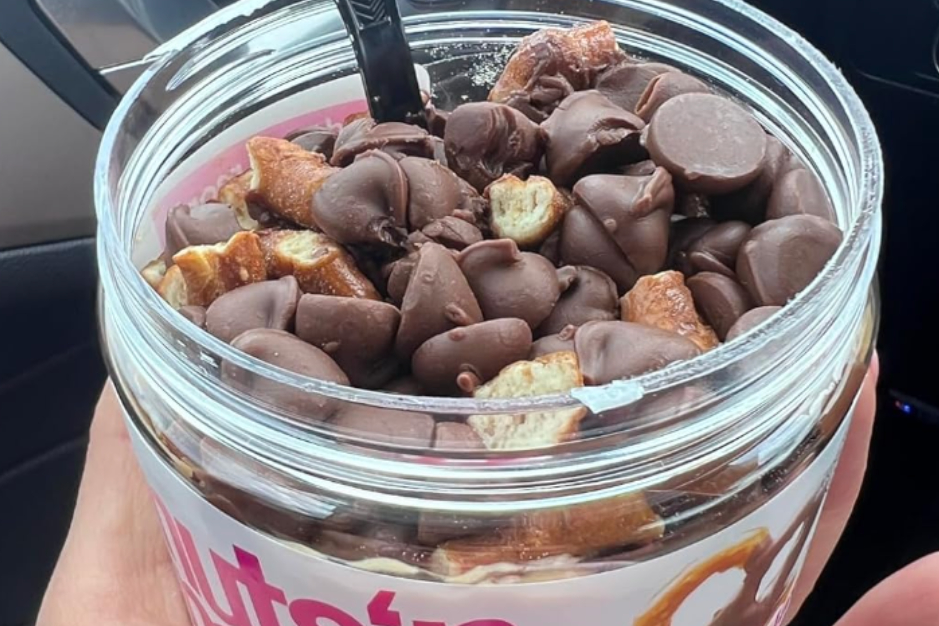 A hand holds a jar of Nuts 'n More, with chocolate-covered pretzel pieces visible on top of the spread