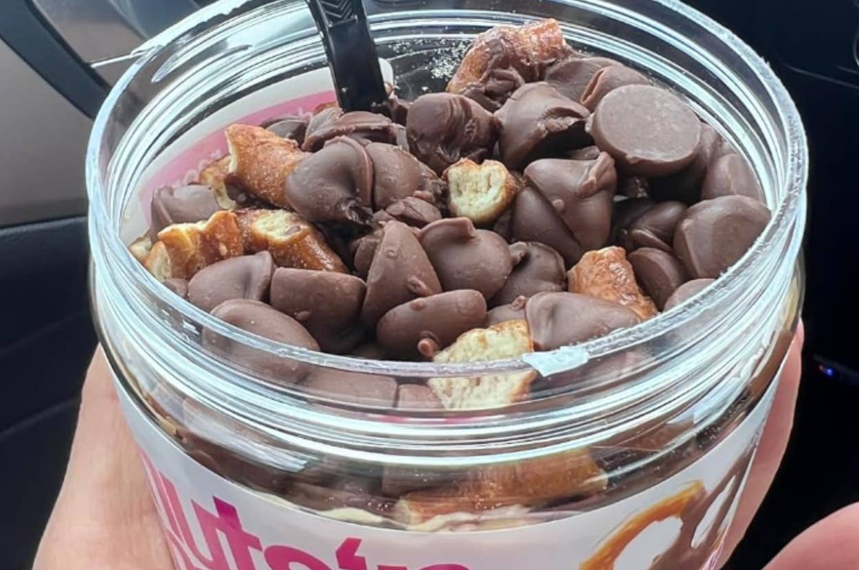 A hand holds a jar of Nuts 'n More, with chocolate-covered pretzel pieces visible on top of the spread