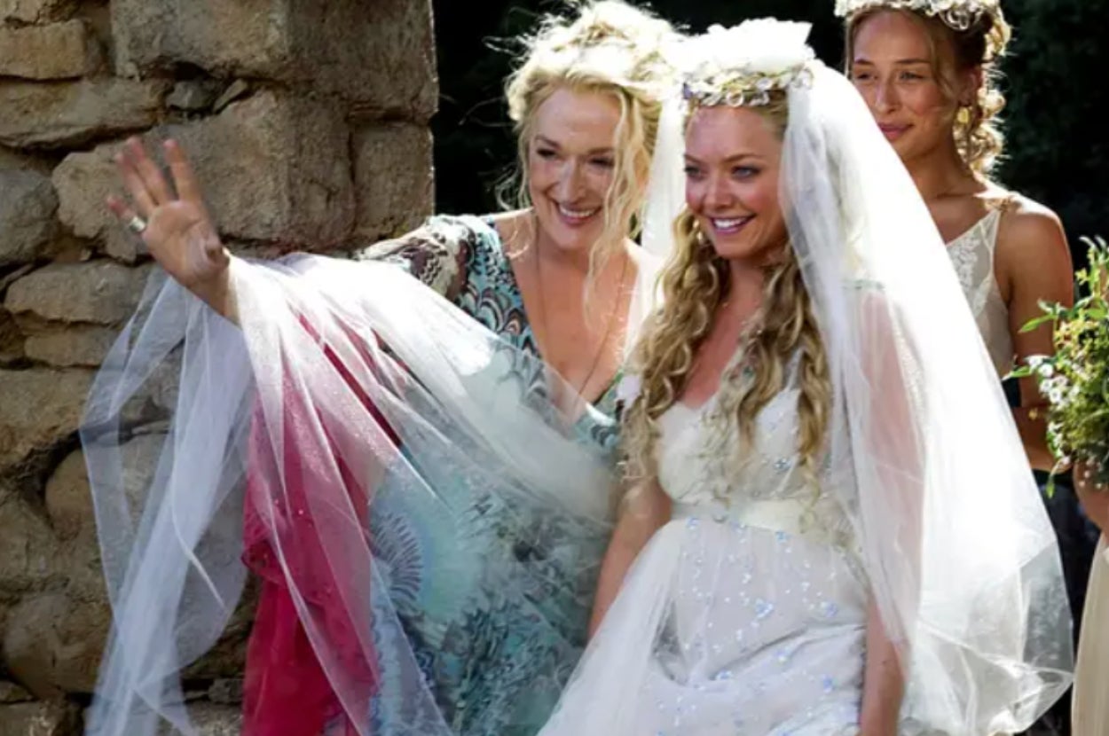 Two women in wedding gowns, both wearing veils and floral headpieces, smiling and waving joyfully near a stone wall, accompanied by a bridesmaid