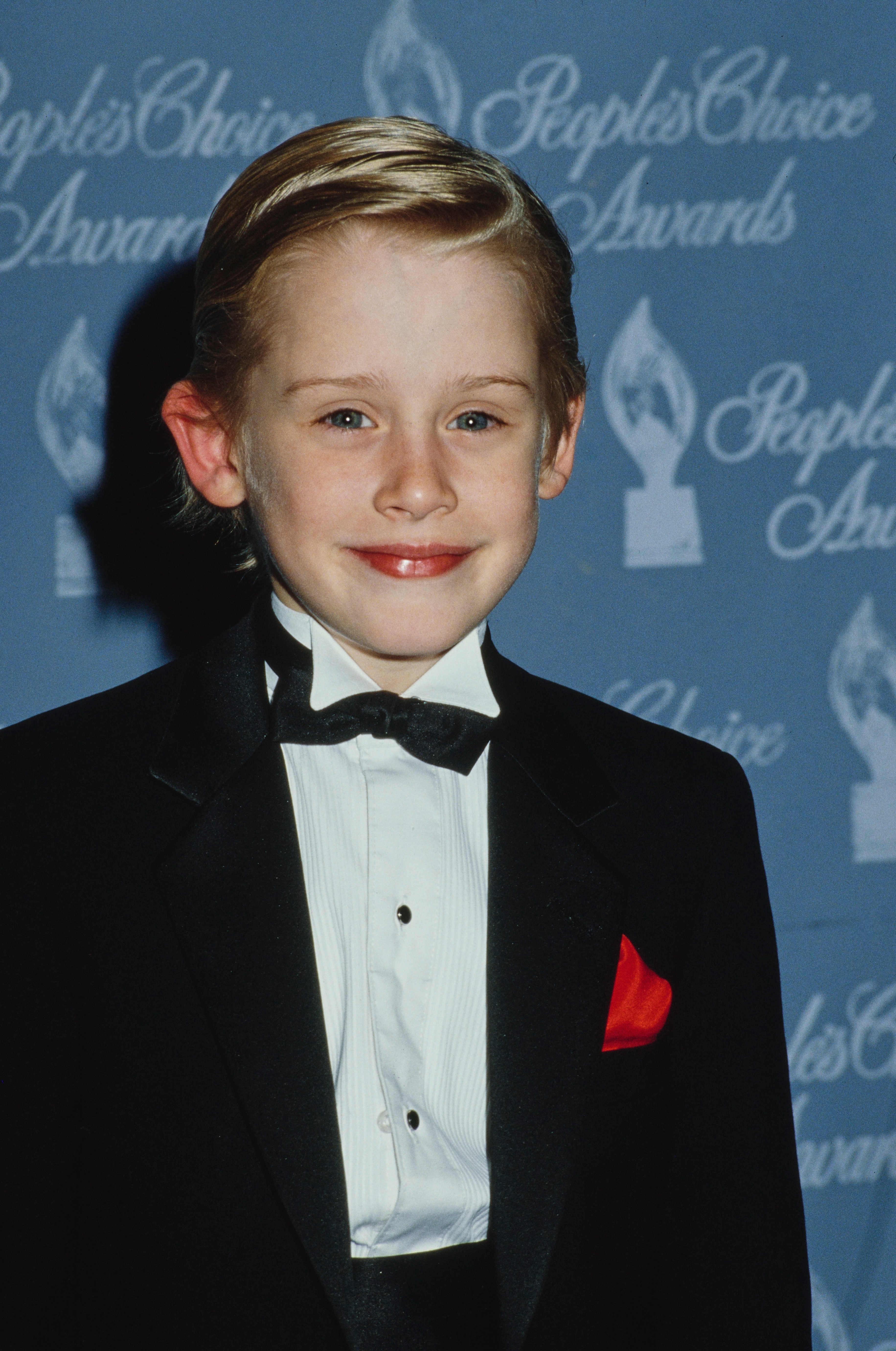 Child histrion  successful  a tuxedo with a reddish  pouch  quadrate  astatine  an awards event, smiling astatine  the camera