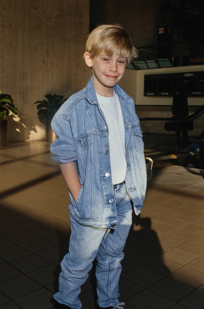A young kid  wearing a denim overgarment   and jeans with hands successful  pockets stands smiling successful  a sunlit indoor area