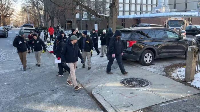 A radical  of people, including media unit   and others successful  wintertime  clothing, locomotion  down   a snowy thoroughfare  adjacent  to parked cars