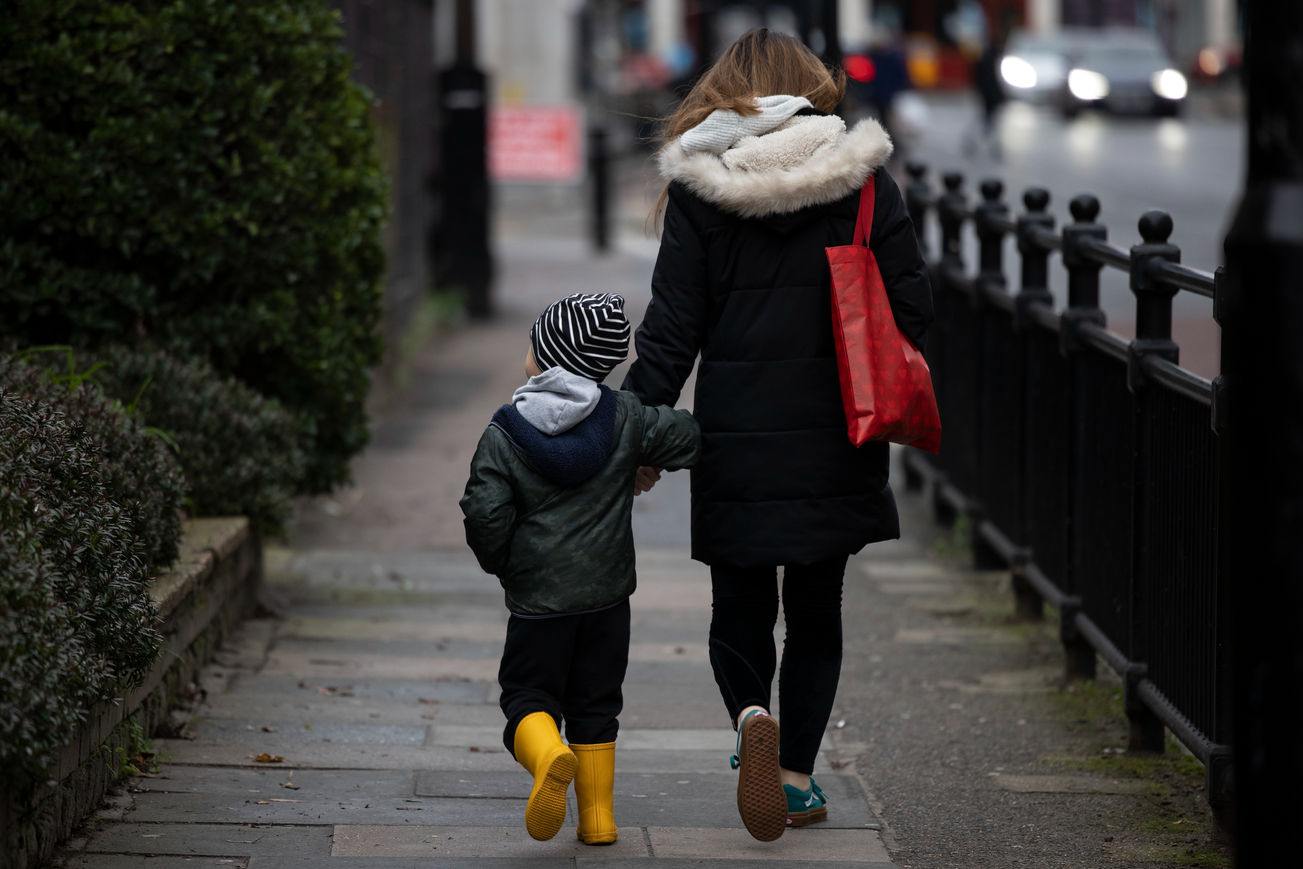 A pistillate and kid locomotion connected a sidewalk holding hands. The kid wears a striped beanie and yellowish boots. The pistillate carries a reddish bag