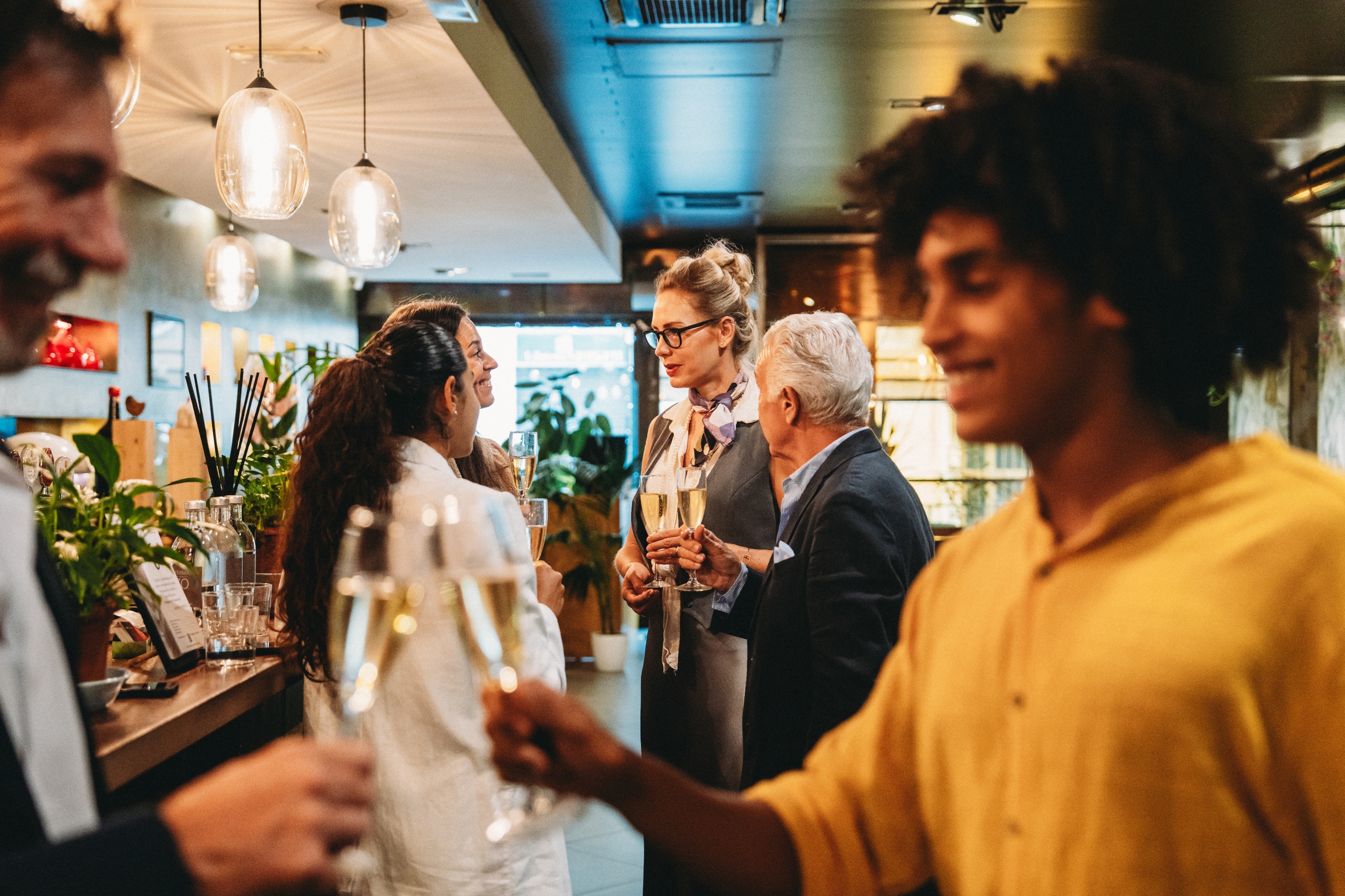 People astatine a societal gathering clinking glasses successful a dimly lit venue. A idiosyncratic successful a yellowish garment is successful the foreground
