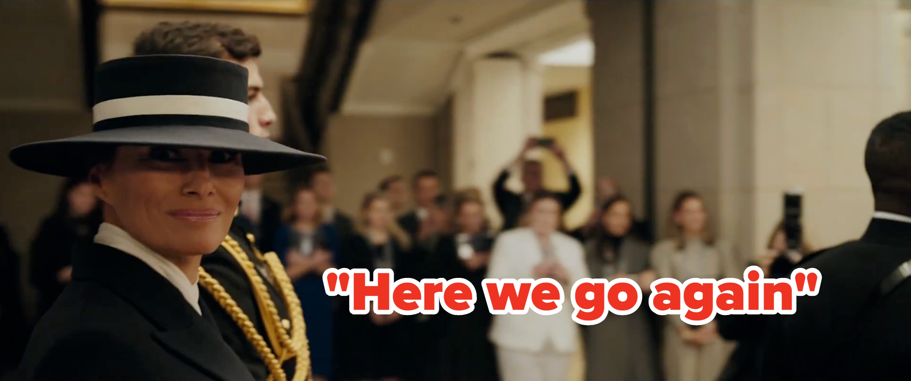 En person i en stilig bredbremmet hatt og skreddersydd dress poserer på et arrangement, deltatt av en folkemengde i formell antrekk i bakgrunnen