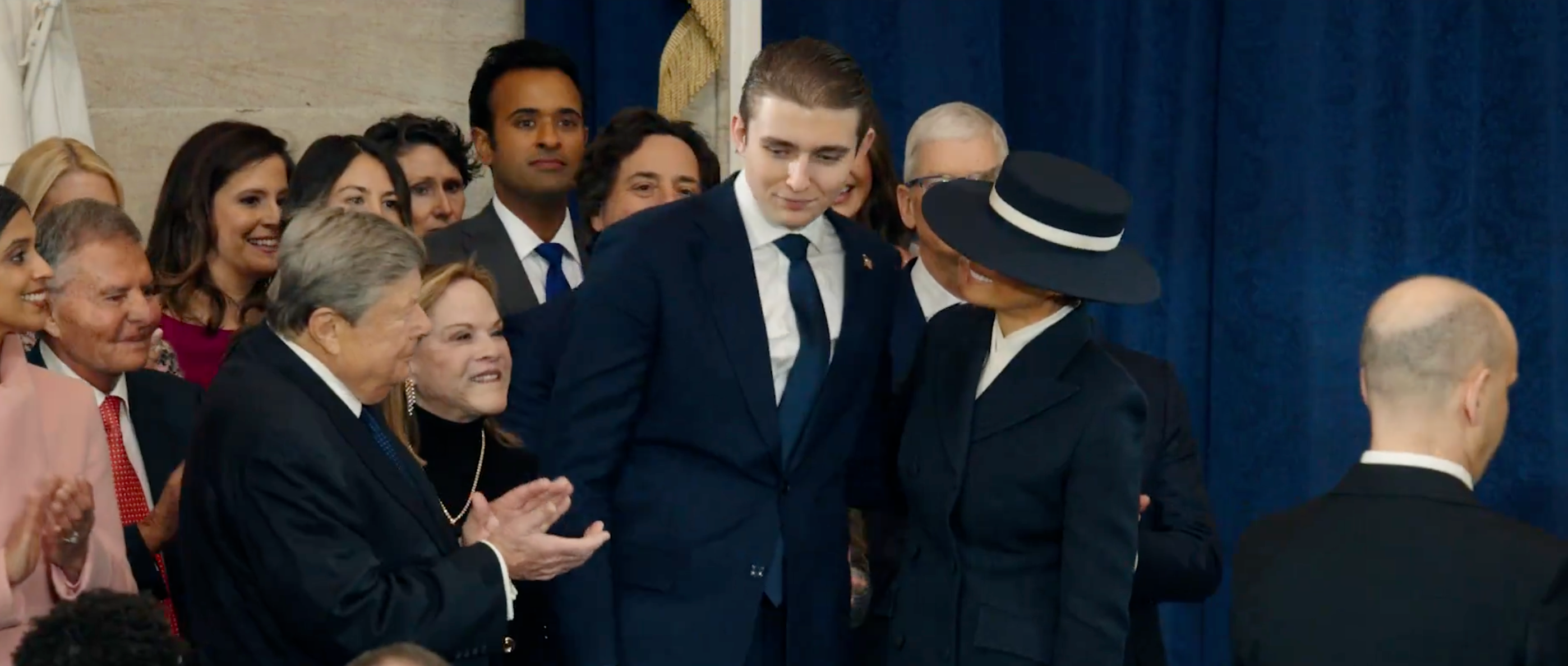 Gruppe mennesker i formell antrekk samlet på et arrangement, en person i dress og en annen i hatt og mørkt antrekk