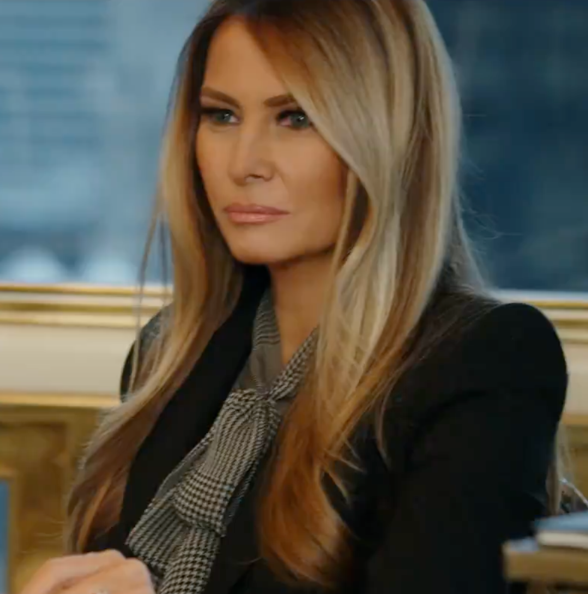 Kvinne med langt hår sitter ved et bord iført forretningsdress med mønstret skjerf