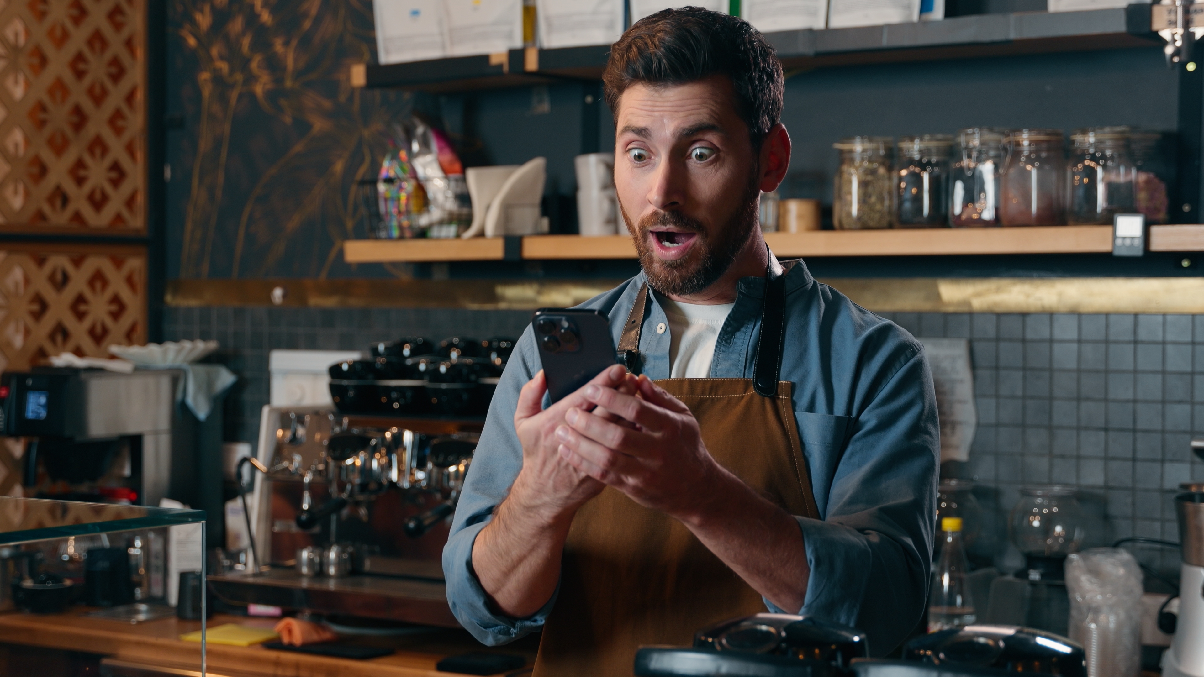 A barista successful an apron looks amazed portion holding a smartphone, lasting wrong a café with java instrumentality around