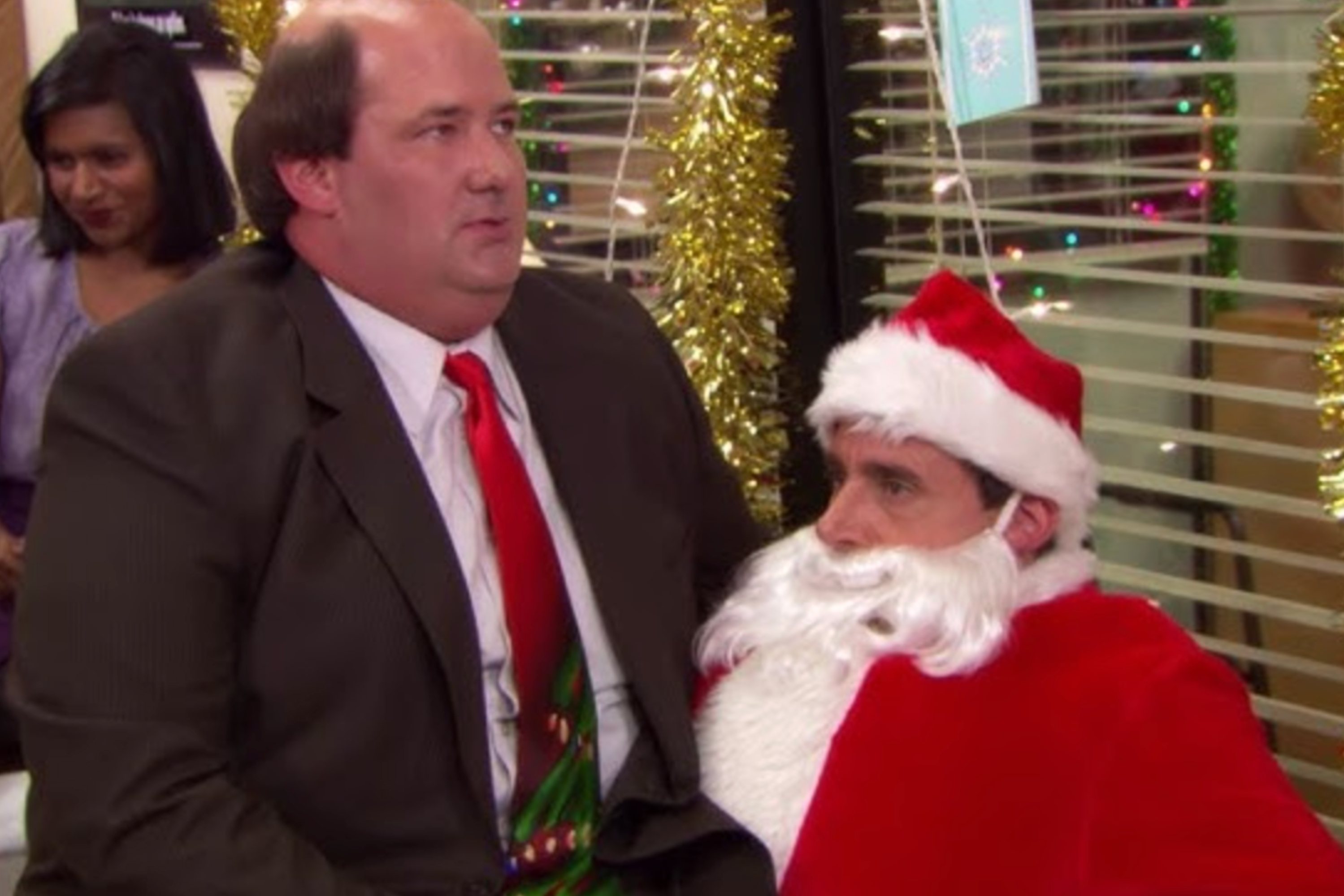 Person in a suit and festive tie sits on a Santa's lap in an office setting, with tinsel decorations in the background