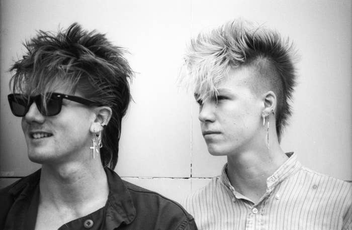 Two radical   with punk stone  hairstyles and earrings. One wears sunglasses and a jacket, portion    the different   dons a striped shirt