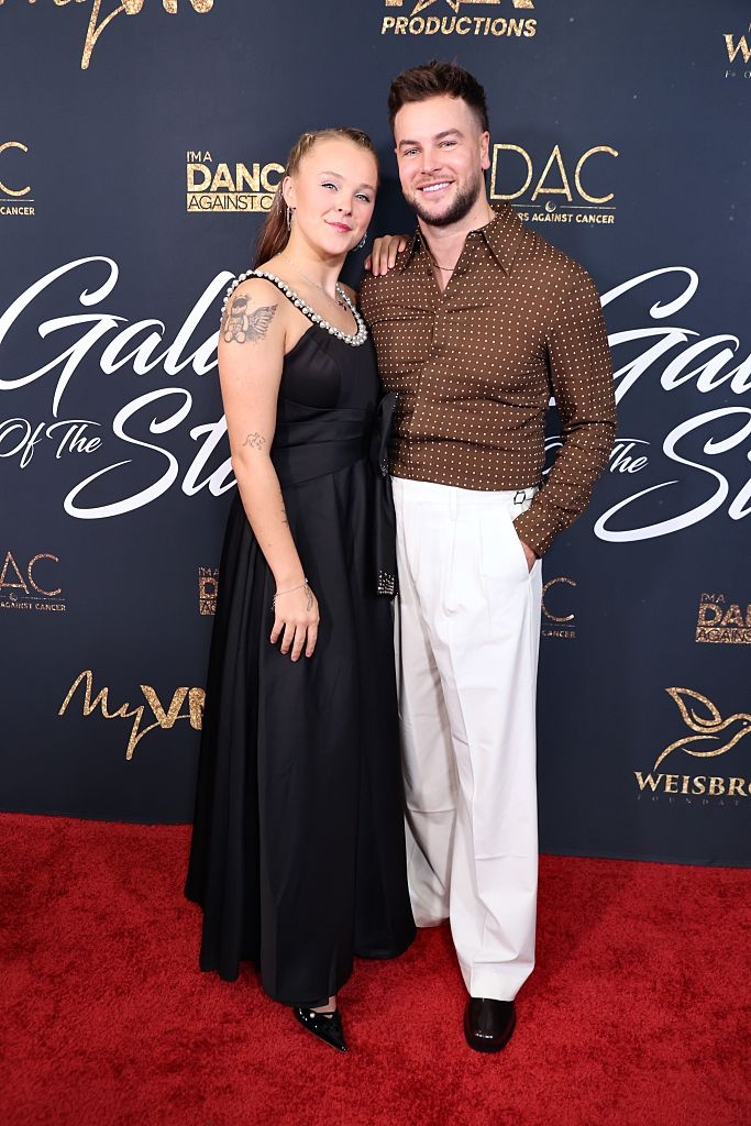 Two radical   astatine  a reddish  carpet event. One wears a sleeveless achromatic  gown with jeweled neckline, the different   a brownish  polka dot garment  with achromatic  pants