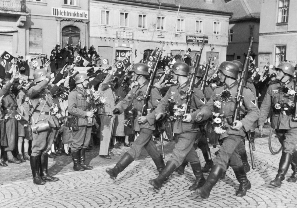 Soldiers successful azygous march successful enactment connected cobblestone street, with onlookers watching from the side. Buildings successful the background
