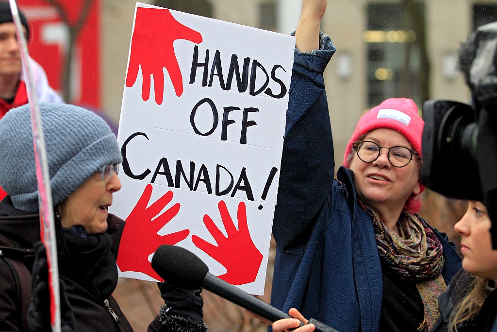 People astatine a protest, 1 holding a motion that reads "HANDS OFF Canada!" with reddish handprints