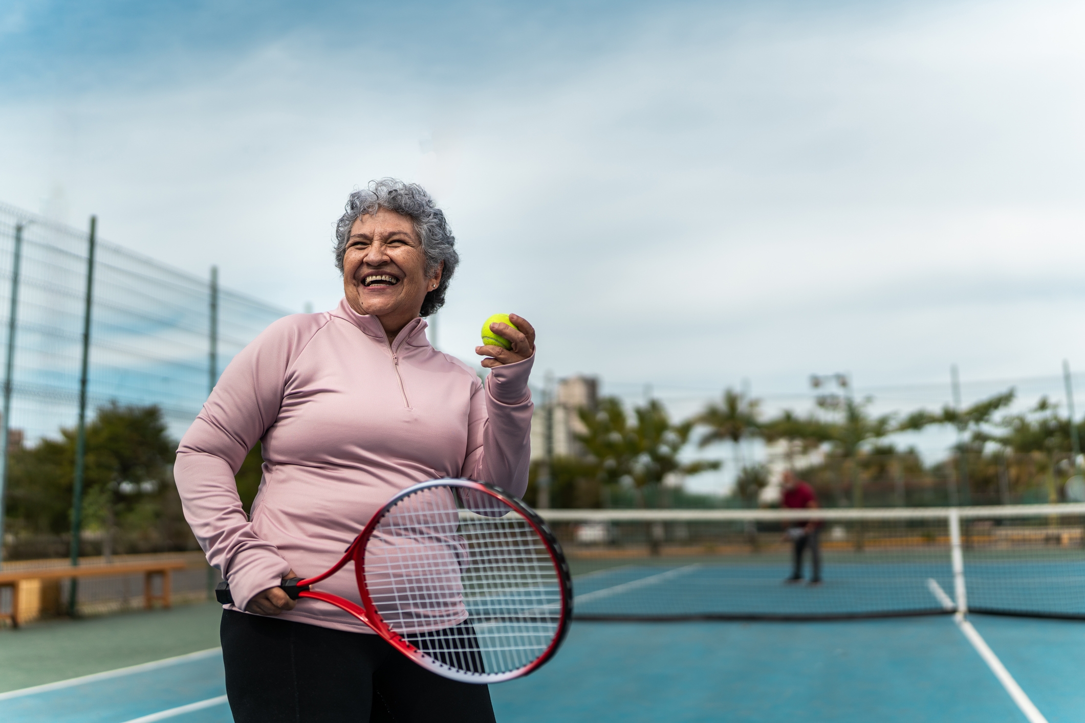Smiling older big  holding a tennis racket and shot  connected  a court, dressed successful  activewear. Another idiosyncratic   is disposable   successful  the background