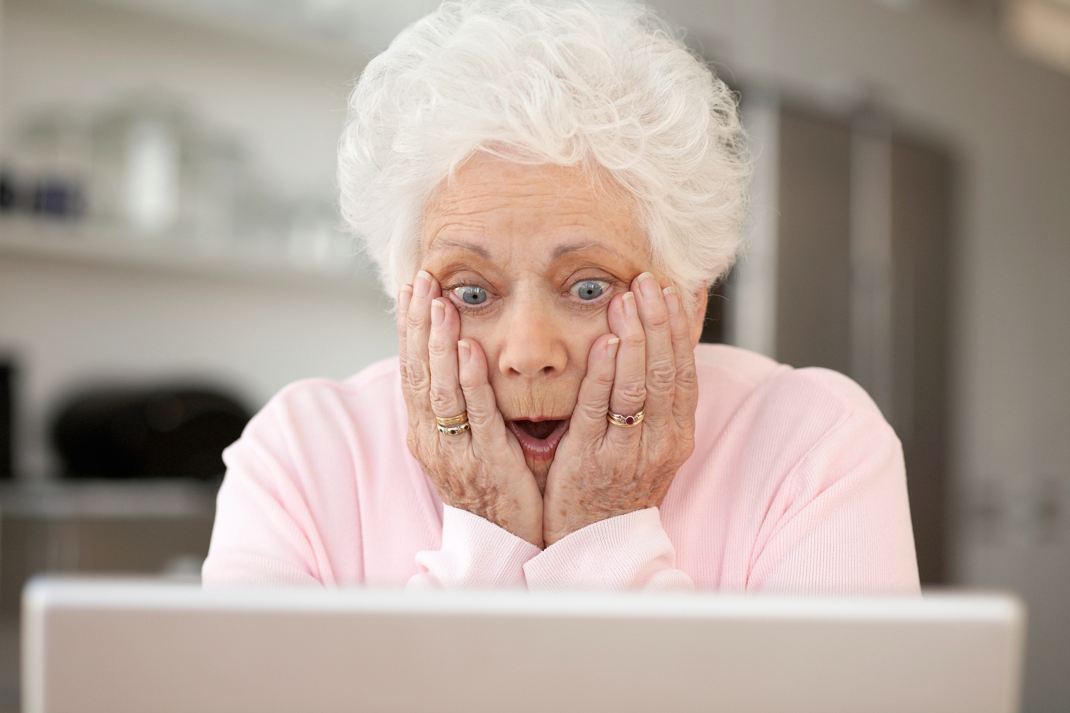 Elderly pistillate   with achromatic  hairsbreadth  and a shocked look  looks astatine  a machine  screen, hands connected  cheeks, seated indoors