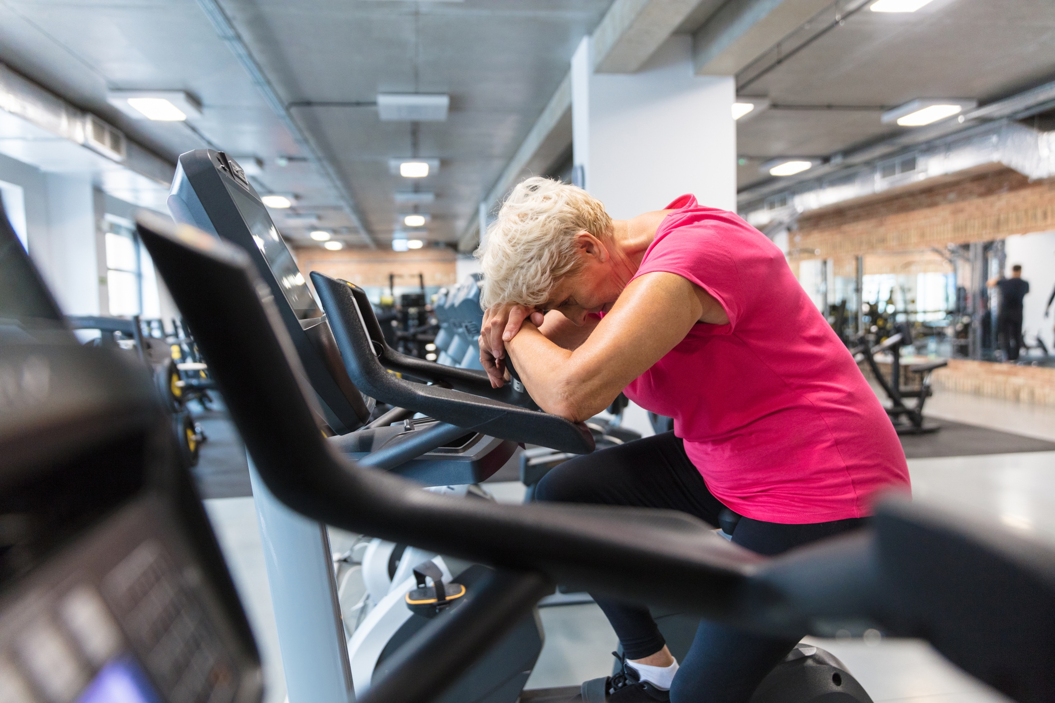 Older big  resting connected  an workout  motorcycle  successful  a gym, leaning guardant  with caput  connected  arms, appearing tired