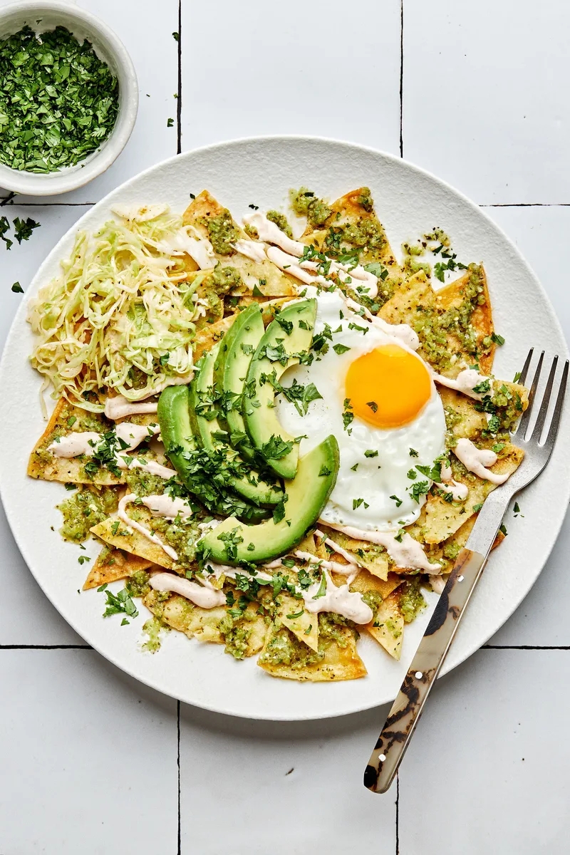 Plate of chilaquiles topped with avocado slices, fried egg, crema, and cilantro, served with shredded cabbage connected  the side