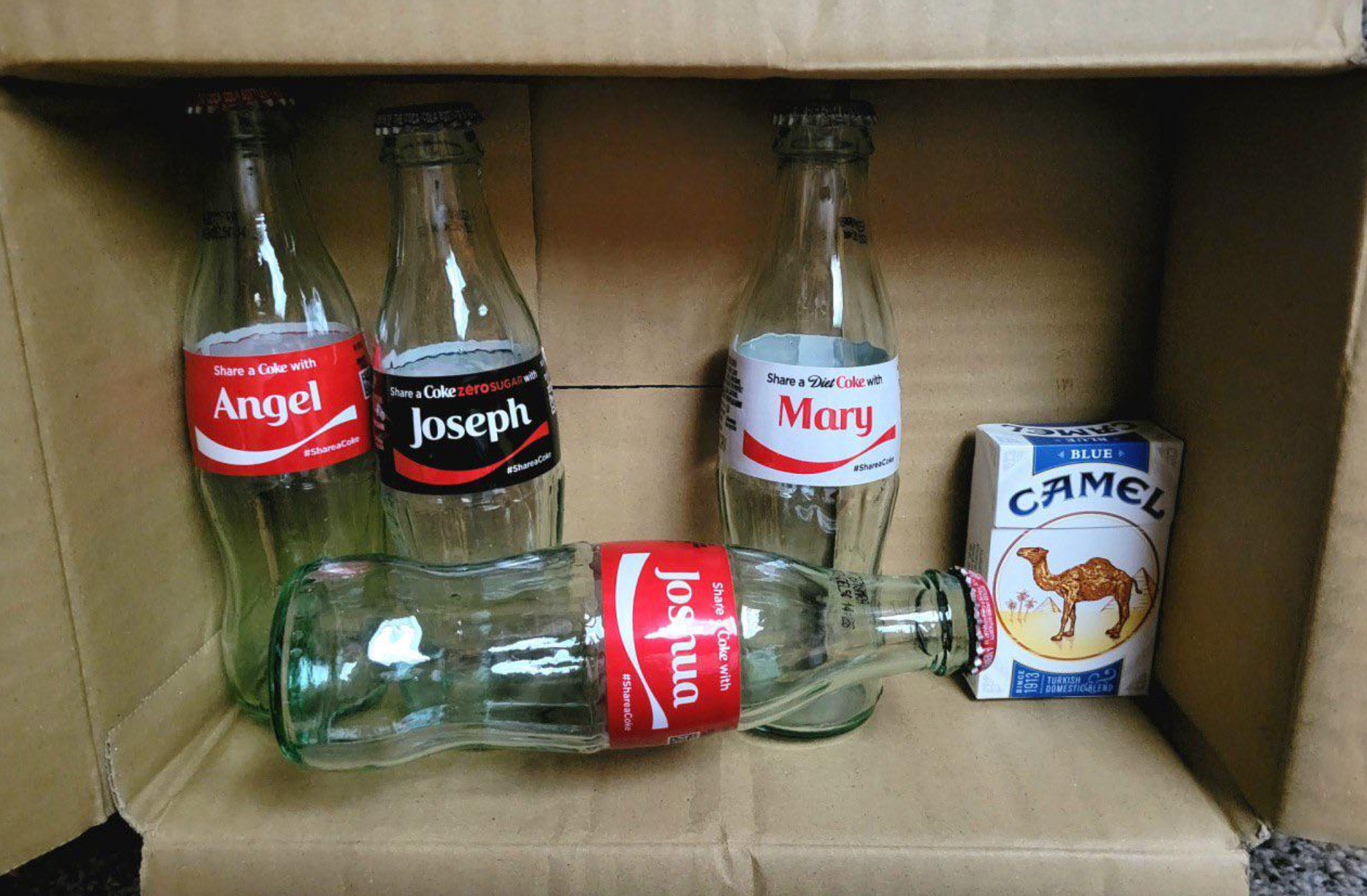 Three soda bottles labeled Angel, Joseph, and Mary basal   upright. A vessel  labeled Joshua lies down. A container  of Camel cigarettes is adjacent  to the bottles