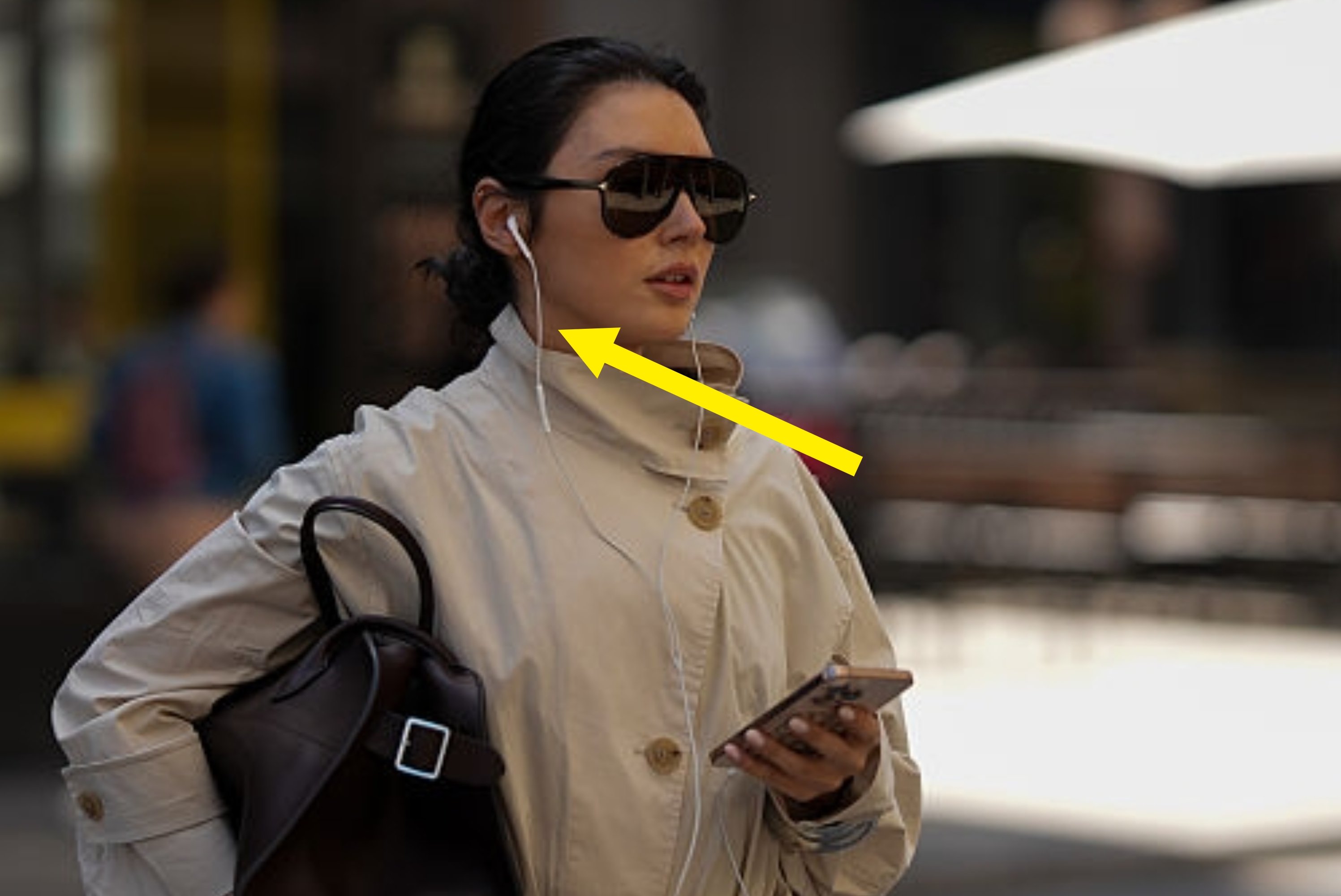 Person successful  a trench coat, sunglasses, and earphones walking connected  the street, holding a telephone  and ample  handbag