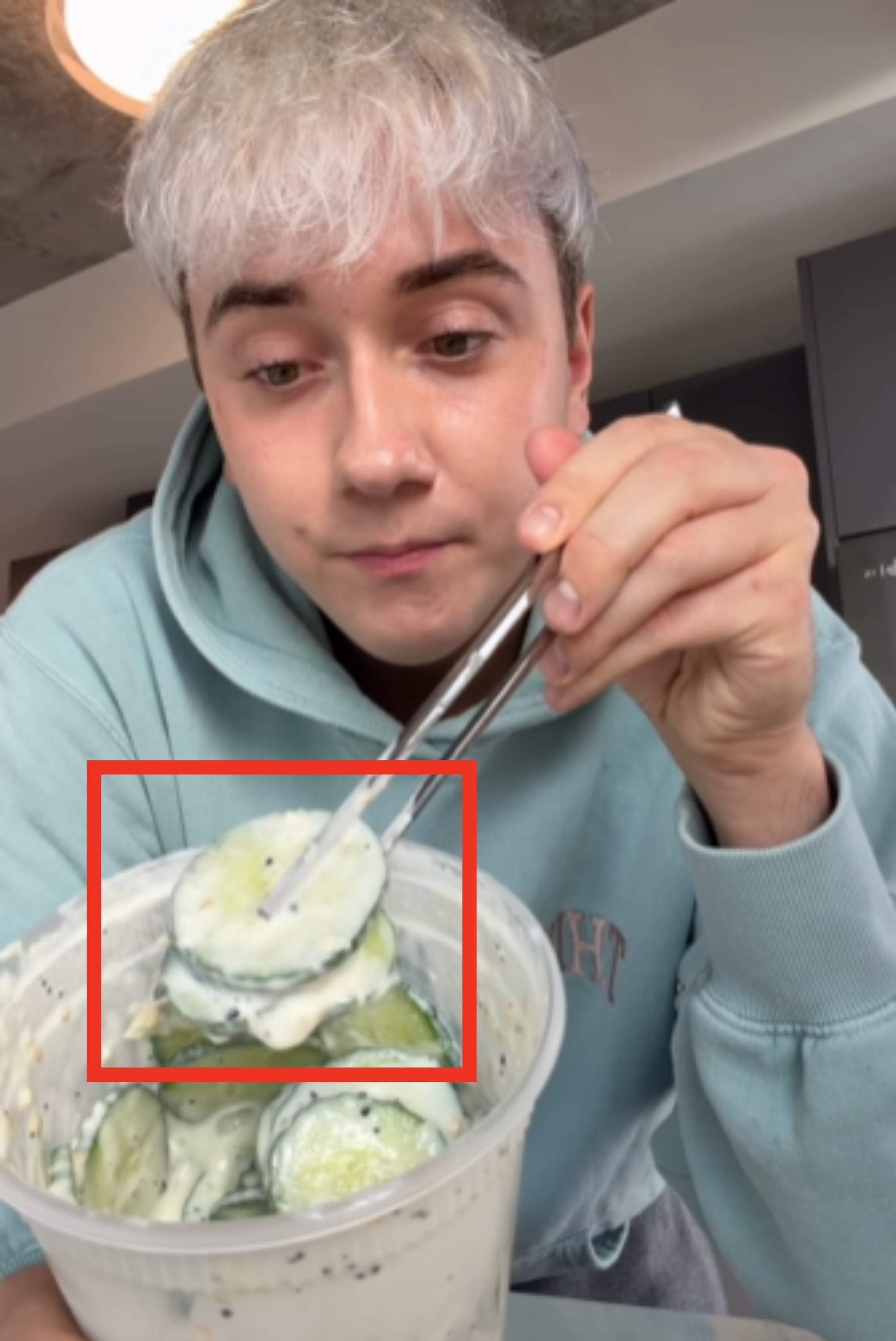 Person successful  casual hoodie holds chopsticks and eats cucumber slices covered successful  a creamy condiment  from a ample  container