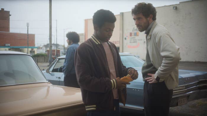 Two radical   basal   by a car   having a conversation, 1  holding an envelope. Another idiosyncratic   is seen successful  the inheritance  adjacent   different  car