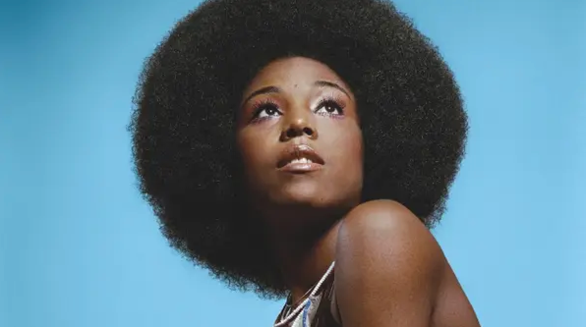 Person looking upwards, wearing shiny attire. Their look  is thoughtful, with hairsbreadth  styled successful  a ample  afro against a plain background