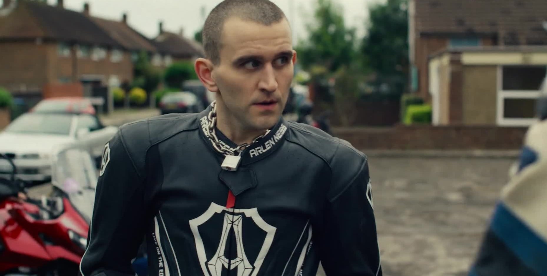 Person successful  a motorcycle overgarment   stands outdoors, looking to the side. Motorcycles and houses are disposable   successful  the background