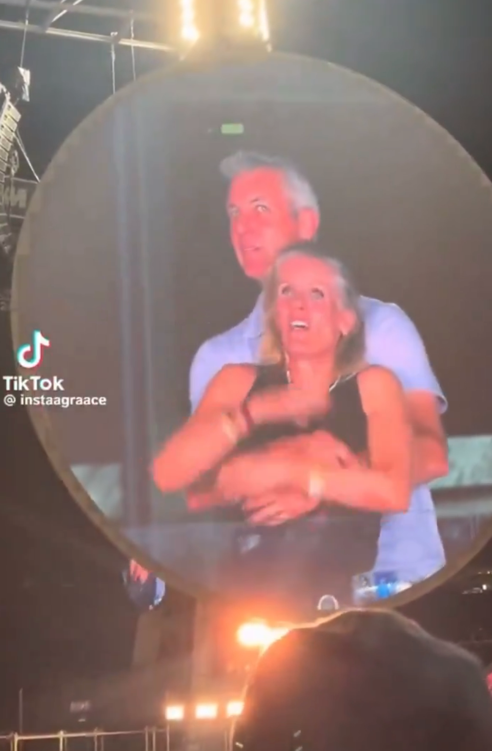 A couple dances together, smiling and embraced, on a large screen at a lively outdoor event